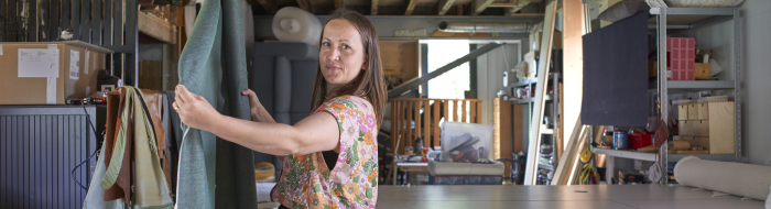 Een vrouw in een stofatelier spreidt een hangende lap stof en kijkt in de camera