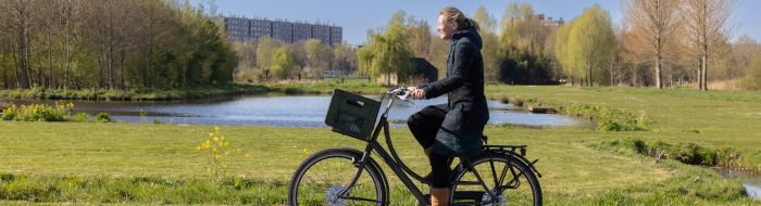 Vrouw fiets door het groen van Noorderpark-Ruigenhoek met flatgebouw op de achtergrond