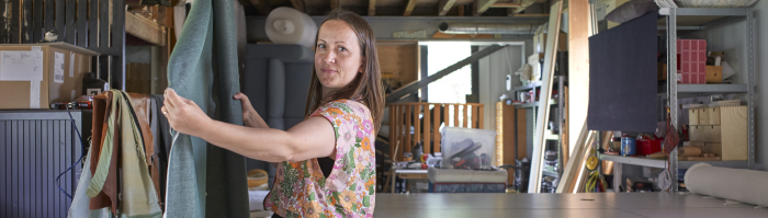 Een vrouw in een stofatelier spreidt een hangende lap stof en kijkt in de camera