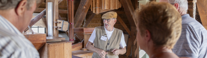Een man geeft aan drie andere mensen een uitleg. Op de achtergrond is het houten interieur van een molen zichtbaar.